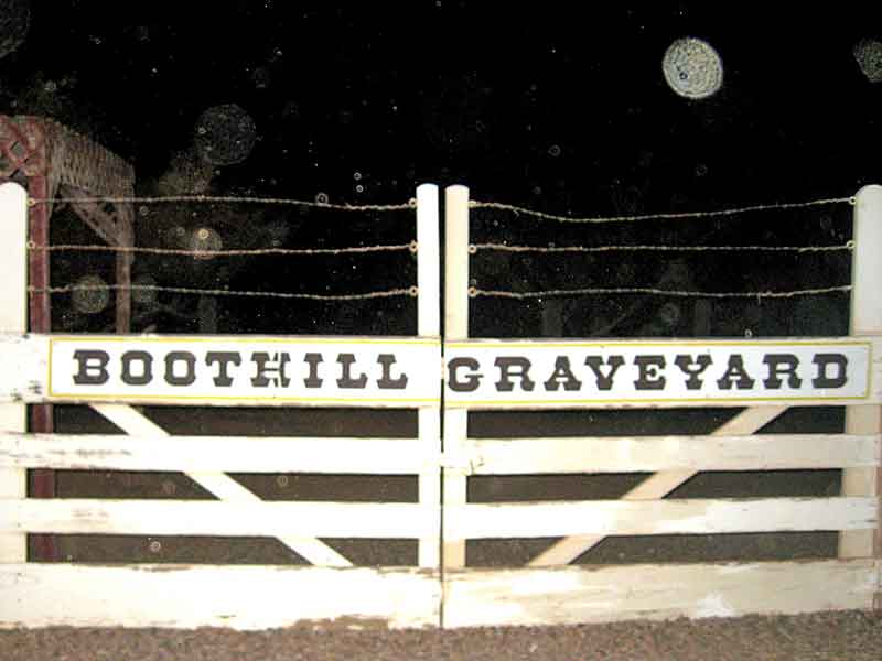 Night photo of the entrance to Boothill Graveyard. A white wooden gate with 'BOOTHILL GRAVEYARD' painted on it is visible. Above the gate, strands of barbed wire can be seen. Several orbs of varying sizes are floating in the dark sky, with one large, distinct orb visible in the upper right corner.