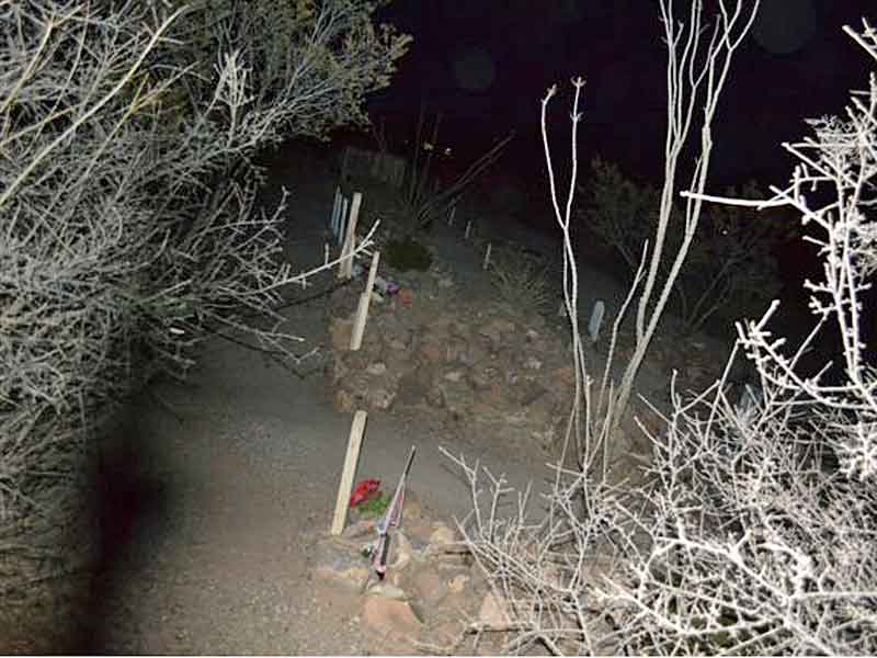 Night photo of Boothill Graveyard. A dirt path winds between graves marked with wooden crosses and stones. Bare, white branches of ocotillo cacti frame the edges of the image. In the foreground, a dark, shadowy shape resembling a human figure can be seen on the left side of the path, suggesting a potential paranormal presence.