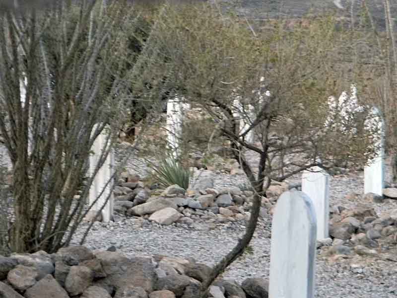 Daytime view of Boothill Graveyard. The rocky, arid landscape is dotted with desert vegetation including ocotillo cacti and small trees. White grave markers are visible among the rocks. In the foreground, a shadowy, translucent figure resembling a human silhouette can be seen walking between the graves, suggesting a paranormal presence.