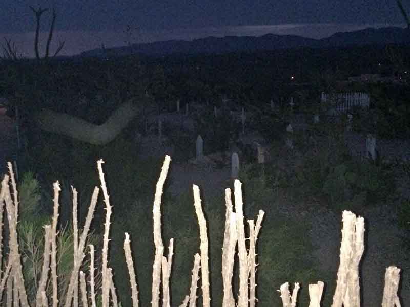 Dusk photo of Boothill Graveyard. In the foreground, pale ocotillo stems form a silhouette. Beyond them, numerous gravestones and crosses are visible in the dimly lit cemetery. Mountains can be seen on the horizon. A distinct, curved, translucent shape resembling a vortex or ectoplasm is visible in the upper left of the image, suggesting paranormal activity.