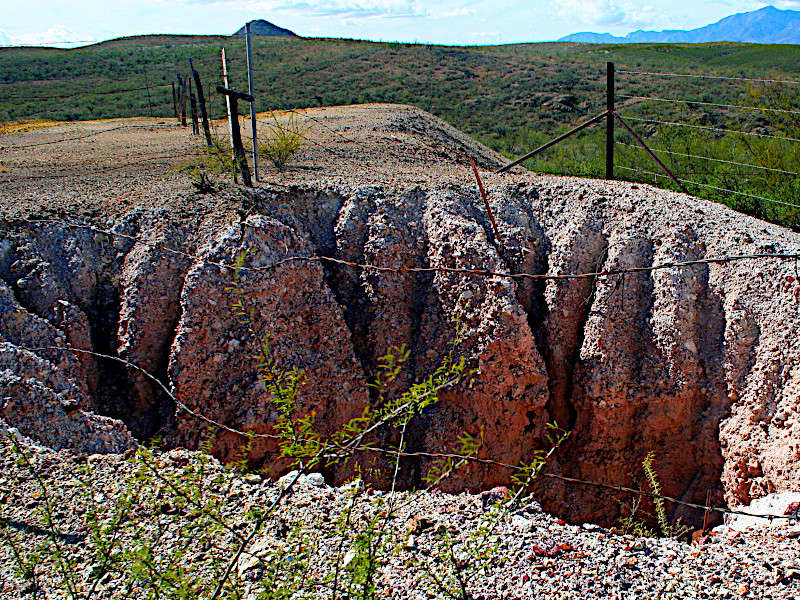 Eroded rocky outcrop with deep grooves, fenced off in a desert landscape. Mountains visible in the background.