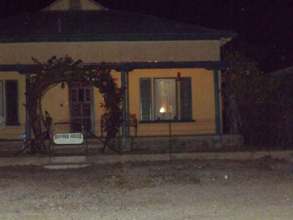 Nighttime view of Buford House, a yellow building with a porch and arched entryway. A sign identifies the house, and a lit window is visible. A ghostly figure is visible at the edge of the porch.