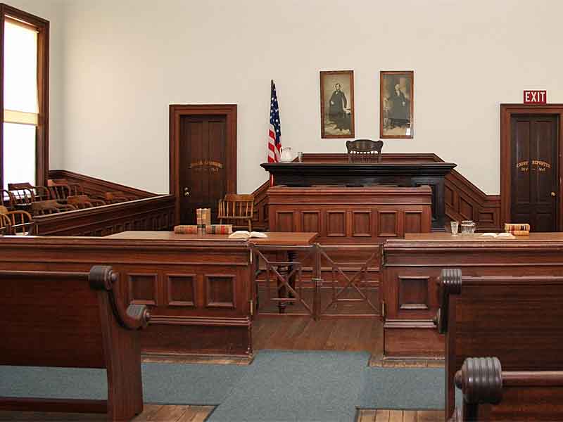 Interior view of the restored Tombstone courthouse courtroom. A judge's bench dominates the front, flanked by two portraits. Wooden jury and spectator seating areas are visible, along with an American flag. The room features rich, dark wood paneling and furnishings, typical of late 19th-century courtrooms.