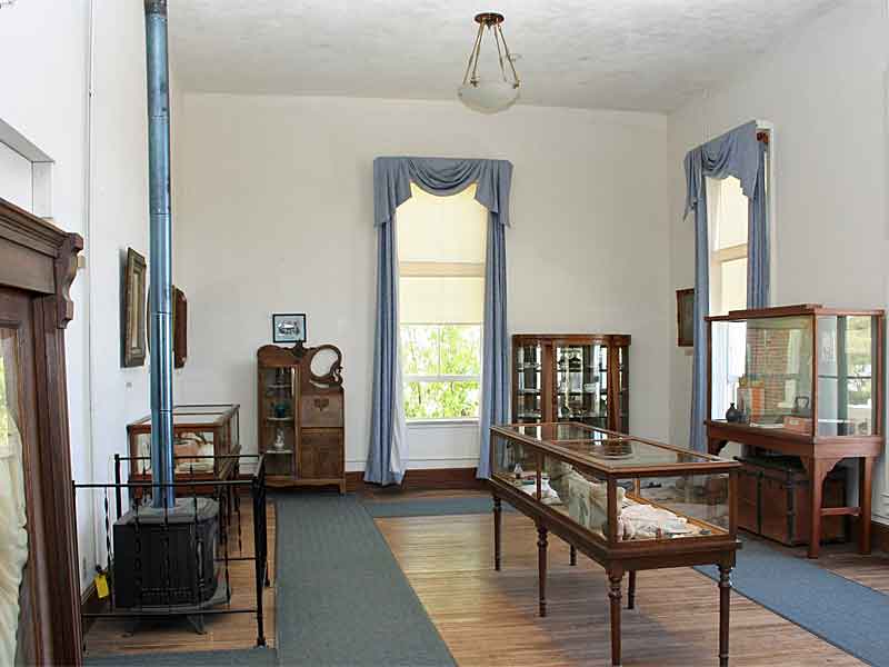 Interior of a room in the Tombstone courthouse museum. The space features period-appropriate decor with tall windows adorned with blue curtains, a vintage light fixture, and wooden floors. Display cases and cabinets showcase historical artifacts. A cast iron stove with a blue pipe stands in the corner, adding to the late 19th-century ambiance.