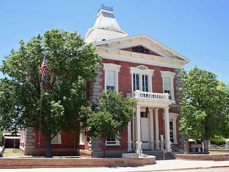 Exterior view of the historic Tombstone Courthouse, now a museum and Arizona state park. The two-story red brick building features white trim, a central balcony, and a distinctive cupola on top. American flags flank the entrance, and mature trees surround the well-preserved structure, highlighting its Victorian-era architecture against a bright blue sky.