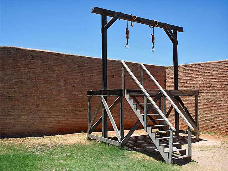 Replica of a gallows in the courtyard of the Tombstone Courthouse Museum. The wooden structure features stairs leading to a platform and a beam with three nooses hanging from it. It stands against a backdrop of high brick walls, illustrating the stark reality of frontier justice in the Old West.