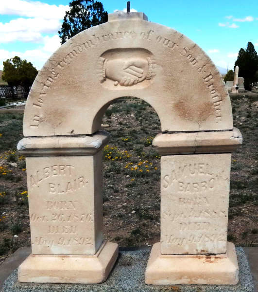 Ornate tombstone arch in a cemetery, with carved text 'In the remembrance of our dear friends' and a handshake symbol. Two pillars support the arch, inscribed with names 'Albert Blair' and 'Samuel Barr' and their respective birth and death dates. Yellow wildflowers bloom in the foreground.