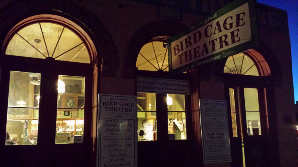 Nighttime exterior view of the Bird Cage Theatre in Tombstone. Illuminated arched windows and a prominent sign above the entrance. The historic building facade is visible with informational placards near the entrance.