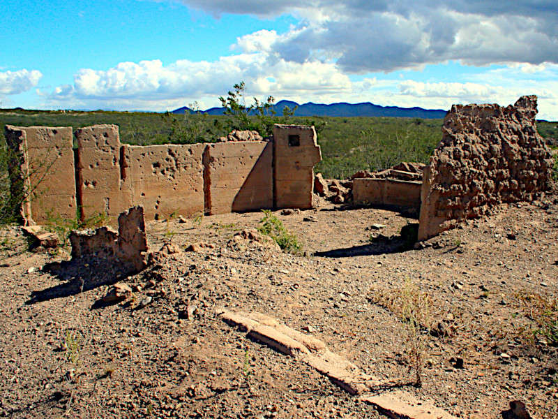 Ruins of an adobe structure in a desert landscape, with crumbling walls and scattered debris. Mountains and cloudy sky visible in the background.