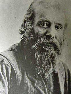 A black and white portrait photograph of an elderly man with a long, flowing beard and intense, penetrating eyes. He has a weathered face with deep wrinkles and is wearing what appears to be a light-colored shirt or tunic. The man's gaze is directed straight at the camera, giving the image a powerful and compelling quality. The portrait captures a sense of wisdom, experience, and strength in the subject's expression and features.