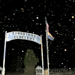 Eerie nighttime view of Tombstone City Cemetery entrance. A white arched sign reading 'City Cemetery' is visible above the gate, with a flagpole to the right. The scene is illuminated by flecks of light, possibly orbs or stars, creating a ghostly atmosphere.