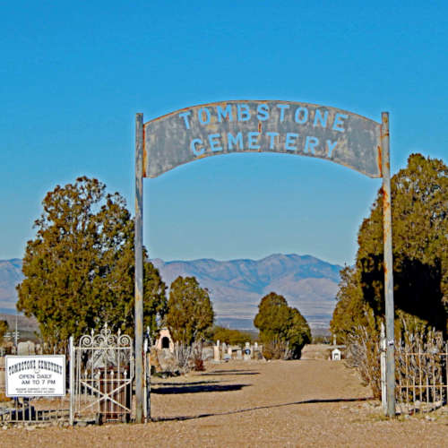 Entrance to Tombstone Cemetery with a weathered metal arch sign. A dirt path leads through an open gate, flanked by cypress trees. In the background, a desert landscape with mountains is visible under a clear blue sky.