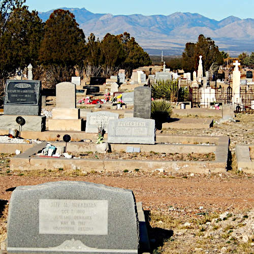 View of Tombstone Cemetery with numerous gravestones and monuments in the foreground. The cemetery is set in a desert landscape with trees and mountains visible in the background. Graves are adorned with various decorations and flowers. The scene captures the historic and rugged atmosphere of the Old West cemetery.