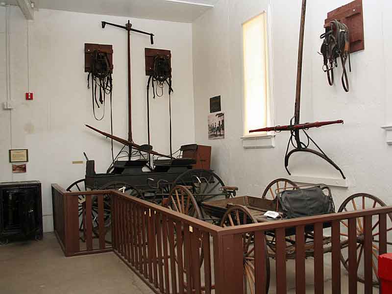 Exhibit at the Tombstone Courthouse Museum showcasing 19th-century transportation. The display features a preserved horse-drawn wagon, various types of horse tack and harnesses mounted on the walls, and other period transportation equipment. A wooden railing surrounds the exhibit, allowing visitors to view the artifacts up close while protecting them.