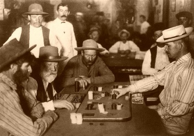 Sepia-toned historical photograph of a crowded saloon in the Old West. Several men are gathered around a gaming table, playing faro or a similar card game. The players and onlookers wear typical late 19th-century attire, including cowboy hats, vests, and button-up shirts. The scene captures the atmosphere of a bustling frontier gambling hall.