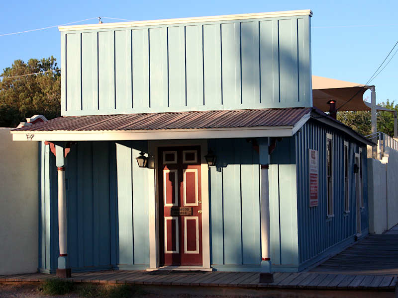 Blue-gray wooden building with a metal roof and red door, typical of Old West architecture. Wooden sidewalk in front and metal awning above.