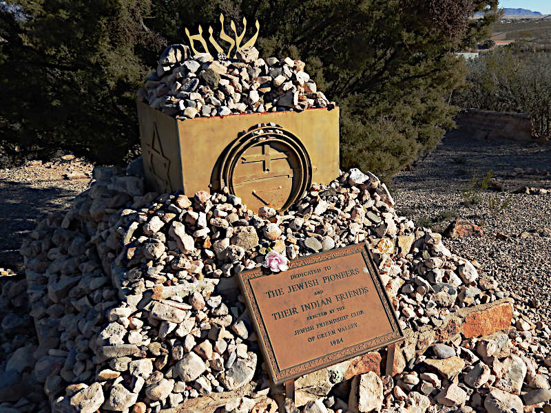 Memorial monument for Jewish pioneers and their Indian friends. A square stone structure filled with rocks, topped with a metal menorah-like design. In front, a pile of stones surrounds a bronze plaque dedicating the monument. The setting is a desert landscape with shrubs and mountains in the background.