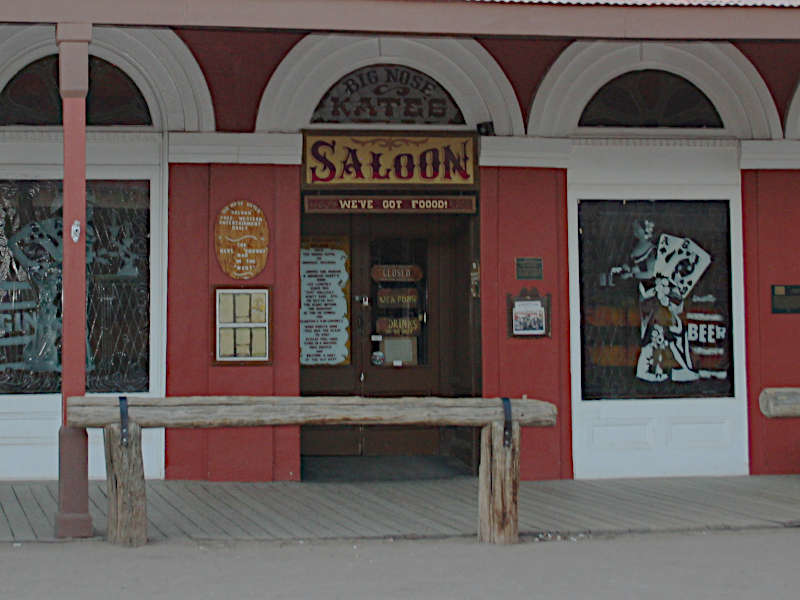 A storefront of an Old West-style saloon with a red exterior and white trim. The entrance features a wooden door with a sign above reading 'SALOON' and 'WE'VE GOT FOOOD!' Arched windows flank the door. A rustic wooden bench sits in front of the building. The windows display various signs and artwork, including a cartoon-style figure holding a beer mug. The facade has an old-fashioned, vintage appearance typical of a Western-themed establishment.