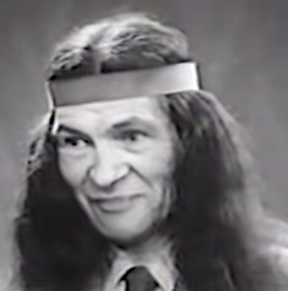 The image shows a black and white portrait of an older man with long dark hair and a headband. He has a friendly smile and is wearing a suit and tie. The man appears to be of Native American descent. The portrait has a soft focus and grainy quality typical of older photographs.