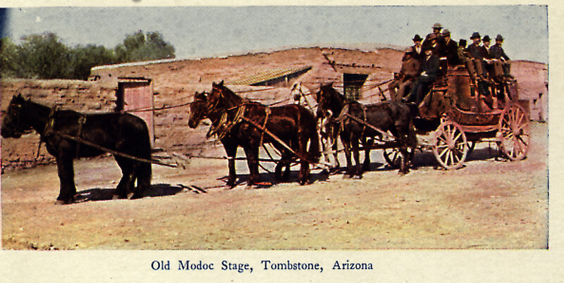 A hand-colored postcard of a Concord stagecoach drawn by a team of dark horses on a dirt road, with a dozen men in hats seated on the roof.