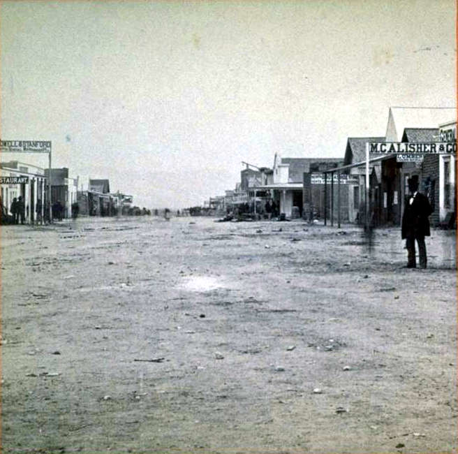 Vintage black and white photograph of a dusty main street in an old western town. Wooden buildings line both sides of the wide, unpaved road. Visible business signs include 'Restaurant' and 'McAlister'. A lone figure in dark clothing stands on the right side of the street. The scene depicts a sparsely populated frontier settlement from the late 19th century.