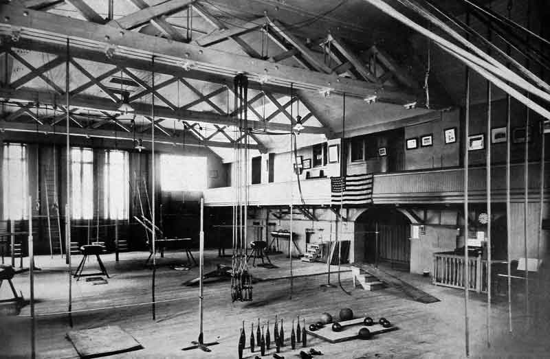 Interior of an old gymnasium with exposed wooden beams and trusses. The room features various exercise equipment including hanging ropes, parallel bars, and Indian clubs on the floor. A balcony or mezzanine level is visible with framed pictures on the wall and an American flag. Large windows on the left provide natural light. The image is in black and white, giving it a vintage appearance.